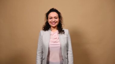 Charming multi-ethnic middle-aged brunette woman, wearing casual suit, waving hello with her hands, greeting and smiling a cheerful toothy smile while looking at camera, isolated on cream background