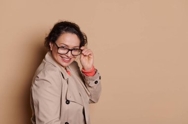 Multi-ethnic mature woman in casual coat, smiling and looking at camera through stylish eyeglasses, isolated over beige background with copy advertising space