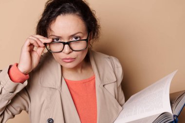 Close-up portrait of a beautiful serious middle-aged dark-haired woman, a professor or teacher holding an open book, looking at camera through her stylish eyeglasses, isolated over beige background