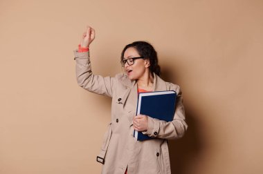 Attractive female professor, a middle-aged dark-haired multi-ethnic woman in stylish beige coat and eyeglasses, having fun, holding a hardcover book, singing and dancing on cream isolated background