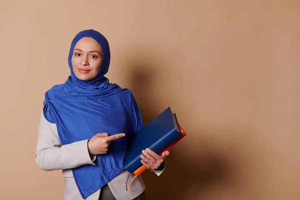 Education. University lifestyle. World book Day concept. Confident Middle-Eastern Muslim female teacher in blue hijab, carrying pile of books, looking at camera, isolated on beige background