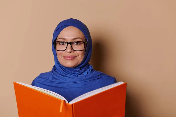 Close-up portrait of a positive smiling Muslim woman with head covered in blue hijab, reads a book with an orange cover and smiles looking at camera, isolated on beige background. People and education