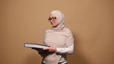 Charming Middle-Eastern Muslim woman wearing casual wear, stylish eyeglasses and head covered in a beige hijab, reading a book with fantasy fairy tails, isolated on cream background