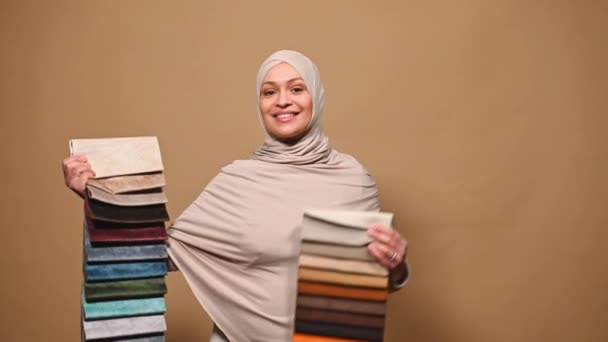 穿着米黄色头巾的阿拉伯穆斯林漂亮女人 惊讶于各种各样的纺织品 推理而不是家庭装饰 选择米色背景的沙发装饰材料 室内设计 — 图库视频影像