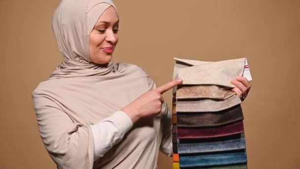 迷人的穆斯林妇女 头戴米黄色头巾 室内设计家或家庭装潢师 向相机展示不同颜色 质地和质量的纺织品样品 用于沙发装饰 — 图库视频影像