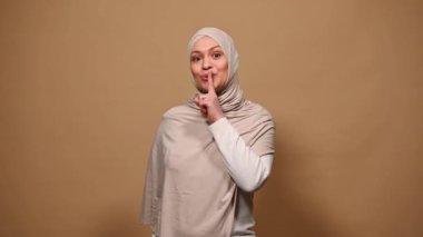 Zevkli bir Arap kadını, şık bir bej elbise ve tesettür giyiyor, el kol hareketi yapıyor, elinde kalp şeklinde pembe bir hediye kutusu, izole bir arka plan tutmadan önce elleriyle sessizlik işareti yapıyor.