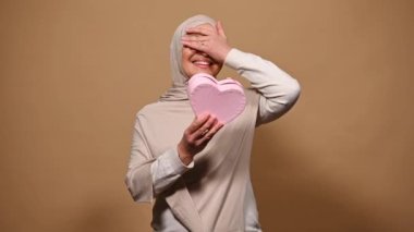 Bej rengi tesettürlü, güzel bir Arap kadın, sürpriz ve olumlu duygularını ifade ediyor, pembe kalp şeklinde hediye kutusunda özel bir hediye kutusunda, izole edilmiş bej arka plan