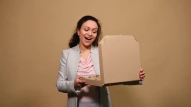 Beautiful happy woman opens a cardboard flat box with hot Italian pizza, shows thumb up and tasty gesture, kisses fingers and says bellissimo, isolated on beige background. Gourmet culinary concept