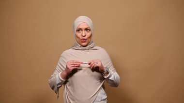 Orta Doğulu, tesettürlü Müslüman bir kadın, hamilelik testi yapıyor, mutluluk ve minnettarlığını dile getiriyor, olumlu sonuçlara bakıyor, bebek beklerken seviniyor, gelecekteki annelik için heyecanlı, izole edilmiş bir zemin