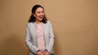 African American happy positive curly-haired middle-aged woman laughing, looking at camera, pointing forefinger at copy space for your advertising text on isolated beige background. Mockup.