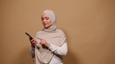 Arap Müslüman kadın tesettürde, akıllı telefondan sosyal medya içeriğini kontrol ediyor, kameraya bakıp gülümsüyor, reklam veya yeni mobil uygulama için fotokopi alanı olan boş siyah ekranı gösteriyor, bej arka plan