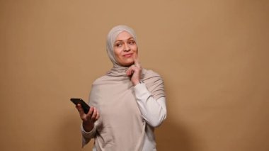 Güzel bir Orta Doğulu Müslüman kadın, bej tesettür takıyor, cep telefonu kullanıyor, arama yapıyor, konuşuyor ve izole bir arkaplanda gülümsüyor. Çevrimiçi iletişim. Tartışma. - İnsanlar. Yaşam biçimi