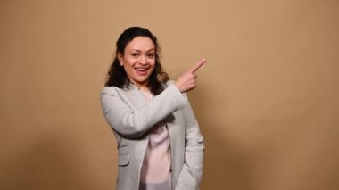 Attractive positive African American middle-aged woman, smiling a toothy smile, gesturing with a thumb up, looking at camera, pointing her index finger at a copy advertising space on beige background