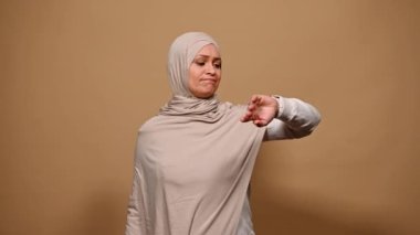 Multitasking Muslim businesswoman checking time on wrist watch, ordering food for lunch time in ecologically-friendly disposable containers, smiling at camera over a beige background. Delivery service