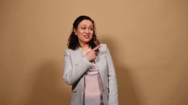 Positive multi-ethnic middle-aged woman, smiling a toothy smile, gesturing with a thumb up, expressing agreement and satisfaction, looking at camera, pointing at copy space for ads on beige background