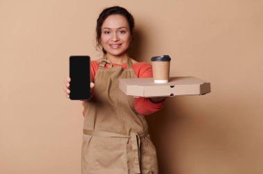 Focus on hands of waitress holding smartphone with blank screen with ad space for mobile apps and cardboard flat box with snacks and disposable paper cup, isolated background. Food delivery service