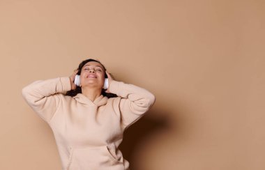 Attractive modern Middle-Eastern woman with closed eyes, wearing wireless headphones, enjoying soothing music, isolated on beige background with copy ad space