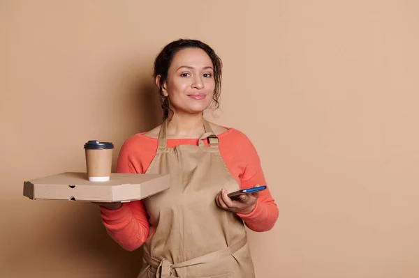 Charming Latin American positive female bartender, waitress in chefs apron, posing with takeaway fast food and mobile phone, isolated on beige backdrop. Online food ordering. Delivery service concept