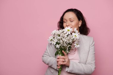 Delighted middle-aged multiracial woman, enjoying smell of a beautiful bouquet of white spring flowers for Womens or Mothers Day, posing with her eyes closed on isolated pink background. Copy space