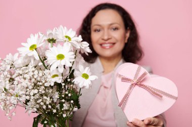 Details on a spring bouquet of white chamomile flowers and gypsophila for Womens or Mothers Day and a heart shaped gift box, in blurred smiling womans hands on isolated pink background. Copy space