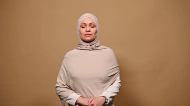 Kendine güveni tam bir Orta Doğulu Müslüman kadın. Başı bej bir başörtüsüyle örtülü, sıkı giyinmiş, kameraya gülümseyen, ellerini çapraz yapan, krem rengi arka planda izole edilmiş.