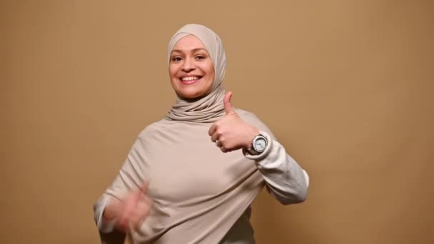 中年正アラブイスラム教徒の女性頭をヒジャーブで覆われました 親指アップ カメラを見て笑顔 肯定的な感情を表現します 同意と孤立ベージュの背景に満足 — ストック動画