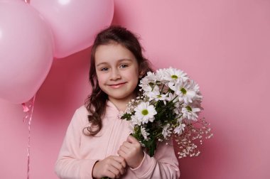 Close-up portrait of Caucasian beautiful little child girl in pink pastel sweatshirt, cutely smiling at camera, posing with a bouquet of white chamomiles on isolated pink background with air balloons