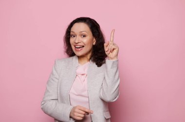 Çok ırklı orta yaşlı çekici bir kadın işaret parmağıyla işaret ediyor, fikri ifade ediyor ve çözüm buluyor, kameraya bakıp gülümsüyor, pembe renkli arka planda izole edilmiş, bedava reklam alanı var.