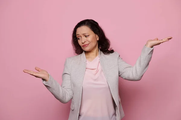 Dark-haired multi-ethnic doubtful woman in formal suit, thoughtfully looking at her hand palm up, weighs the pros and cons of her decision, isolated on pink background with copy space for your ads