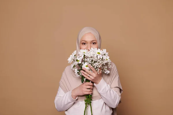 stock image Charming Middle-Eastern Muslim woman with head covered in beige hijab, holding bouquet of white flowers covering half of her face, looking at camera with insightful gaze over isolated cream background