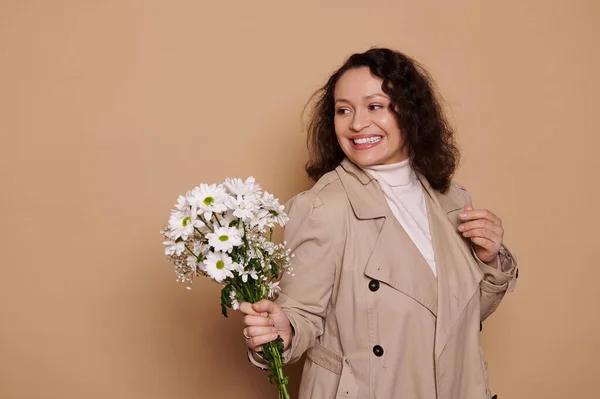 stock image Natural beauty middle-aged woman, wearing a beige coat, holding a bouquet of chamomiles, smiling a beautiful toothy smile looking aside at copy space on isolated background. International Womens Day