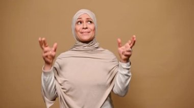 Bej tesettürlü Müslüman bir kadın, dua eden umut dolu bakışlarla, sevgiyle dilek diliyor, hayallerinin gerçekleşmesini umuyor, şans veya iyi şans diliyor, izole bir arka plan diliyor. - İnsanlar. Duygular