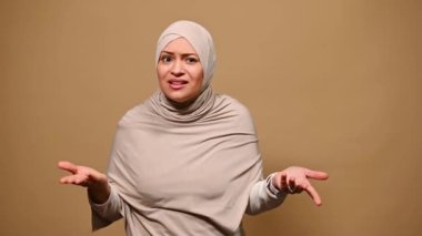 Müslüman bir kadın bej bir başörtüsü takıyor, hayal kırıklığını ve yanlış anlaşılmayı ifade ediyor, ne soruyor, kameraya bakıyor, kendini işaret ediyor ve reddediyor, bej arka planda izole edilmiş.