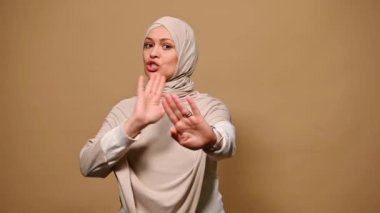 Bej rengi sıkı giysiler ve tesettüre bürünmüş, kendinden emin bir Arap kadın, elleriyle yaptığı jestler, inkar, reddetme ve anlaşmazlığı ifade eden, kameraya bakan, izole edilmiş bir arka plan gösteriyor.