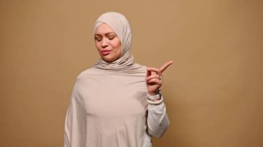 Orta yaşlı, kendinden emin, bej tesettürlü Müslüman kadın, DUR işareti gösteriyor, inkar ediyor, reddediyor ve anlaşmazlık gösteriyor, kameraya bakıyor, izole edilmiş bir arka plan. Kadın hakları kavramı