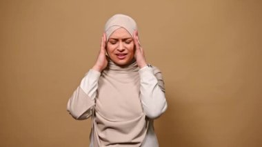 Orta yaşlı, güzel bir Arap kadın bej tesettür takıyor, baş ağrısı çekiyor, çaresiz ve stresli, çünkü acı ve migren, ellerini baş şakaklarına koyuyor, izole edilmiş arka planda.