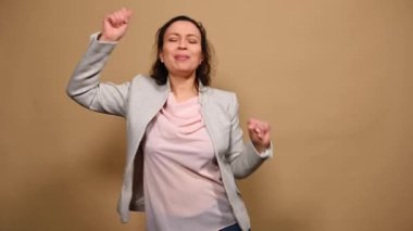 Mutlu çok ırklı orta yaşlı kadın şarkı söylüyor, dans ediyor, müzik dinliyor, neşeli olumlu duygular ifade ediyor, kameraya kurnazca gülümsüyor, izole edilmiş bej arka planda.