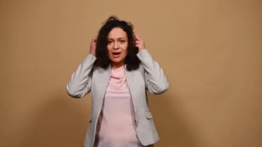 Çok ırklı orta yaşlı kıvırcık kadın, kablosuz kulaklıkla havalı bir parça dinliyor, melodiden hoşlanıyor ve mikrofona girmiş gibi saç fırçasına şarkı söylüyor, dans ediyor ve izole edilmiş bej arka planda hareket ediyor.
