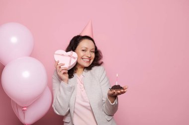 Güzel bir kadın, neşeli bir şapka takıyor, elinde güzel bir kalp şeklinde hediye kutusu ve doğum günü hediyesi var, kameraya gülümsüyor, yanan mum ve balonla zehirliyor, izole edilmiş bir arka planı var.