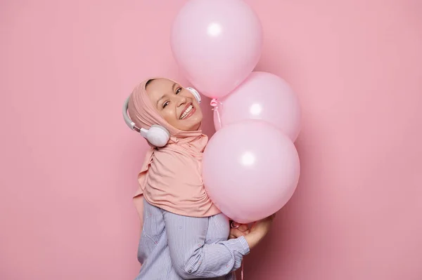Attractive glamour Middle-Eastern Muslim woman in a pink hijab and wireless headphones, having fun on her birthday party, listens to music, smiles, poses with pastel color balloons on pink background