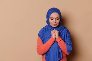 Kafası mavi tesettüre bulanmış Orta Doğulu Müslüman bir kadın ellerini göğüs hizasında tutarak umut dolu, bej arka planda izole edilmiş bir şekilde barış için dua ediyor. İnsanlar ve Din. Boşluğu kopyala