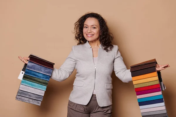 Pleasant curly-haired multi-ethnic woman, interior designer or sales manager in furniture store, showing you a palette of upholstery fabric swatches, smiling at camera on isolated beige background