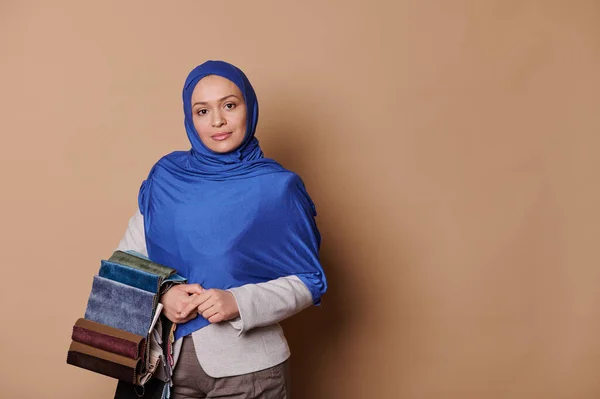 Professional portrait of a Middle-Eastern Muslim woman in formal clothes and blue hijab, interior designer holding upholstery fabric samples, looking confidently at camera, isolated beige background