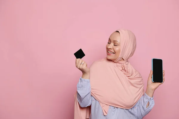 stock image Smiling Middle-Eastern Muslim pretty woman in pink hijab, holding credit card and showing at camera a smartphone with blank black screen for your ads or mobile applications, isolated pink background