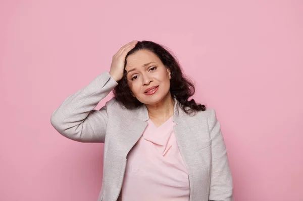 Overworked multitasking woman, businesswoman, manager suffers from tension headache, holding hand on her head, looking at camera with desperate look and hopeless, isolated over pink color background