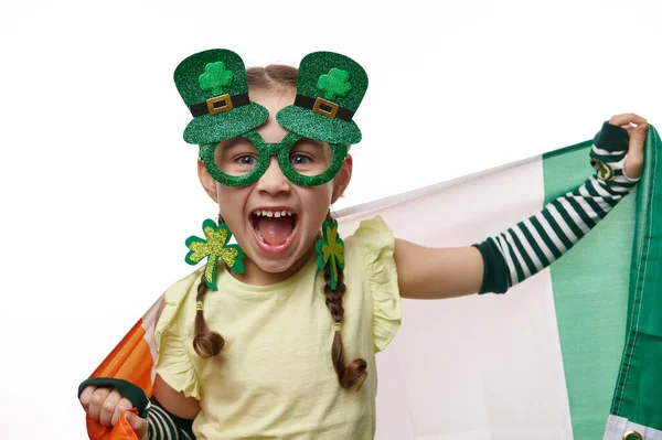 淘气的爱尔兰小女孩穿着时髦的嘉年华服装 穿着Leprechaun的衣服 在镜头前做鬼脸 拿着爱尔兰国旗 被白色背景隔离 圣帕特里克节的概念 — 图库照片