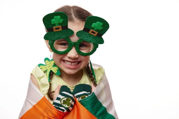 特写可爱可爱可爱的爱尔兰小女孩 戴着时尚的狂欢节护目镜和三叶草耳环 带着可爱的微笑 头戴爱尔兰国旗 背景为白色 圣帕特里克节 — 图库照片
