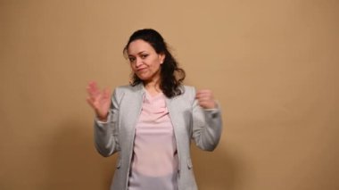 Saldırgan, öfkeli, çok ırklı kadın, günlük kıyafetler içinde, ilk el hareketleriyle avuç içini tekmeliyor, tehdit işaretini elleriyle gösteriyor, kameraya bakıyor, bej arka planda tek başına duruyor.