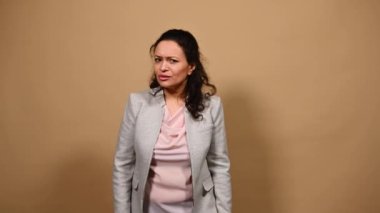Öfkeli bir kadın, kameraya bakarak, başparmakları aşağıda el kol hareketi yaparak, hoşnutsuzlukla bakarak, şaşkınlığını dışa vurarak, izole edilmiş bej arka planda şaşkınlığını ifade ederek, derin nefes alarak,