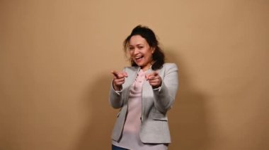 Güzel, orta yaşlı, çok ırklı bir kadın, sıradan bir ceket giymiş, gülüyor, gülümsüyor ve seni işaret ediyor, bej rengi arka planda soyutlanmış.